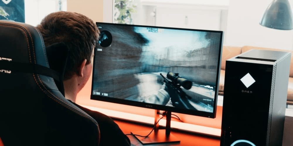 A gamer wearing headphones playing a first-person shooter game on a monitor next to an OMEN gaming PC tower with LED lighting.